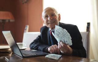 Happy senior businessman holding money in hand while working on laptop at table